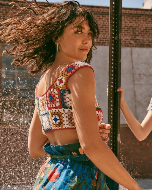 Brunette woman in multicolored crochet crop top and patterned skirt smiling outdoors, sunlight on her hair, urban environment in background.