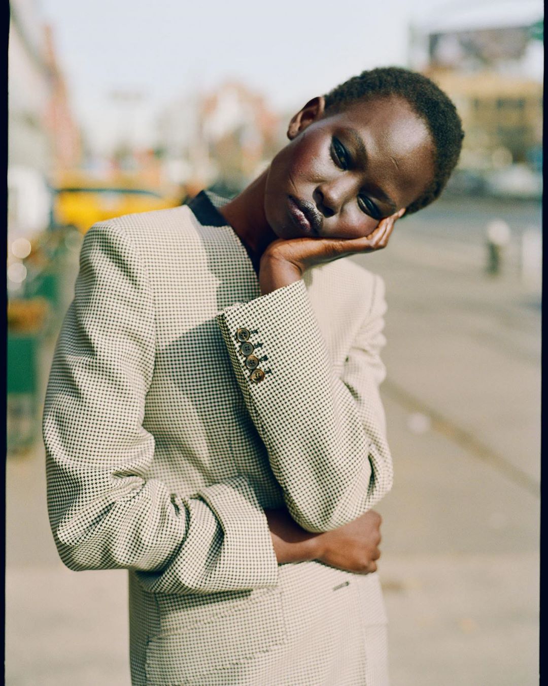 Adult woman with short dark hair poses in a patterned beige jacket outdoors. Her hand supports her head, which is tilted slightly.