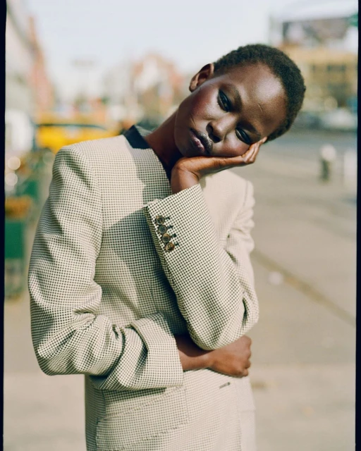 Adult woman with short dark hair poses in a patterned beige jacket outdoors. Her hand supports her head, which is tilted slightly.