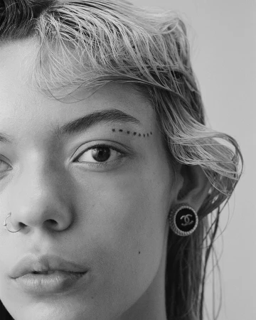 Close-up portrait of a woman with wet hair, nose ring, eyebrow dots, and Chanel logo earrings on a neutral backdrop.