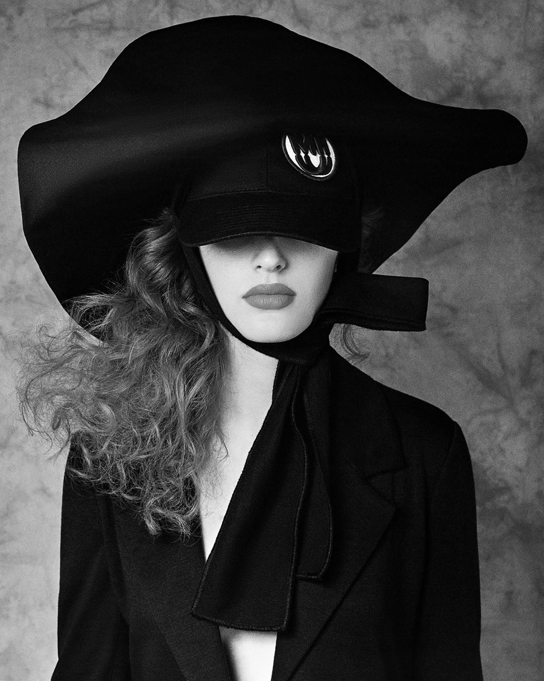 Black and white portrait of a woman wearing a large hat with a visor and an open black jacket against a textured background.