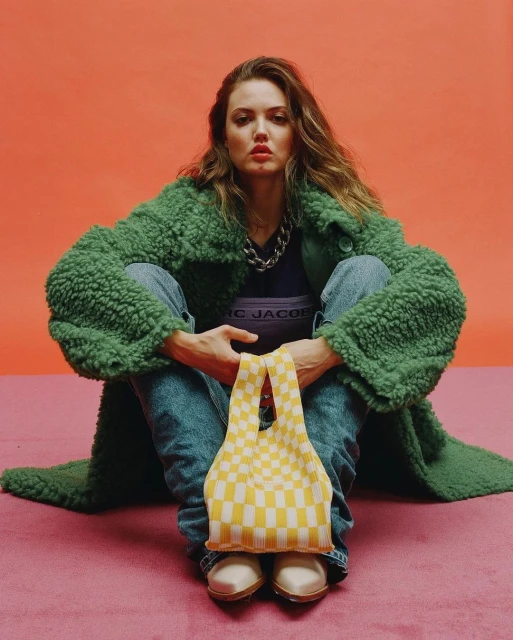Woman in a green coat and jeans sitting on a pink surface, holding a yellow-and-white checkered bag; an orange background.