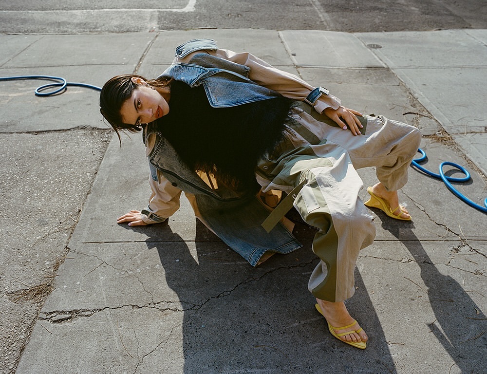 Woman in denim jacket and cargo pants crouching on a concrete sidewalk by a blue hose, photographed by Tiffany Nicholson.