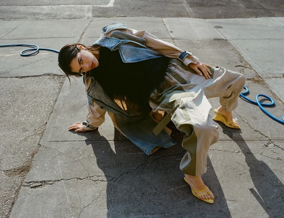Woman in denim jacket and cargo pants crouching on a concrete sidewalk by a blue hose, photographed by Tiffany Nicholson.