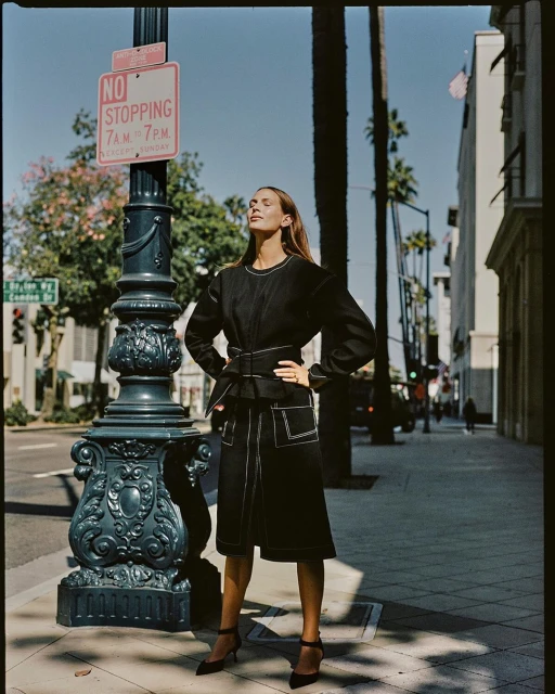 A woman in a black skirt suit stands on a sidewalk next to a black lamp post under a 'No Stopping' sign, bathed in sunlight.