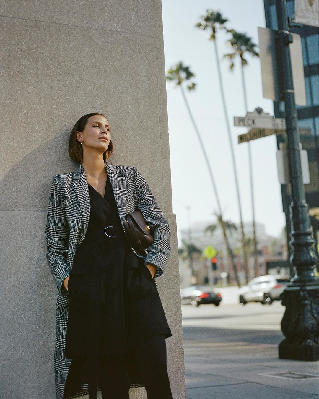 A woman poses against a wall outdoors in a black dress and patterned overcoat, holding a purse, with palm trees in the background.