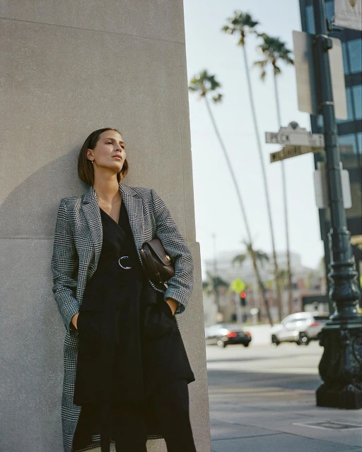 A woman poses against a wall outdoors in a black dress and patterned overcoat, holding a purse, with palm trees in the background.