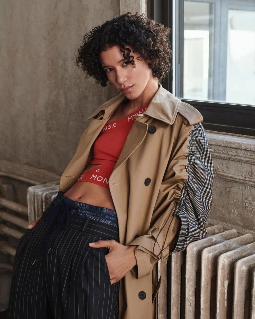 Fashionable young woman with curly hair poses in a studio wearing a red crop top, wide-legged striped pants, and a tan trench coat.