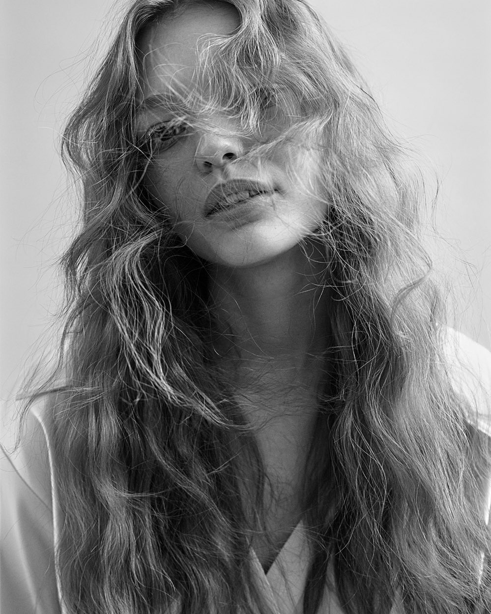 Black and white portrait of a young adult woman with long, wavy hair partially covering her face, wearing a light-colored shirt.