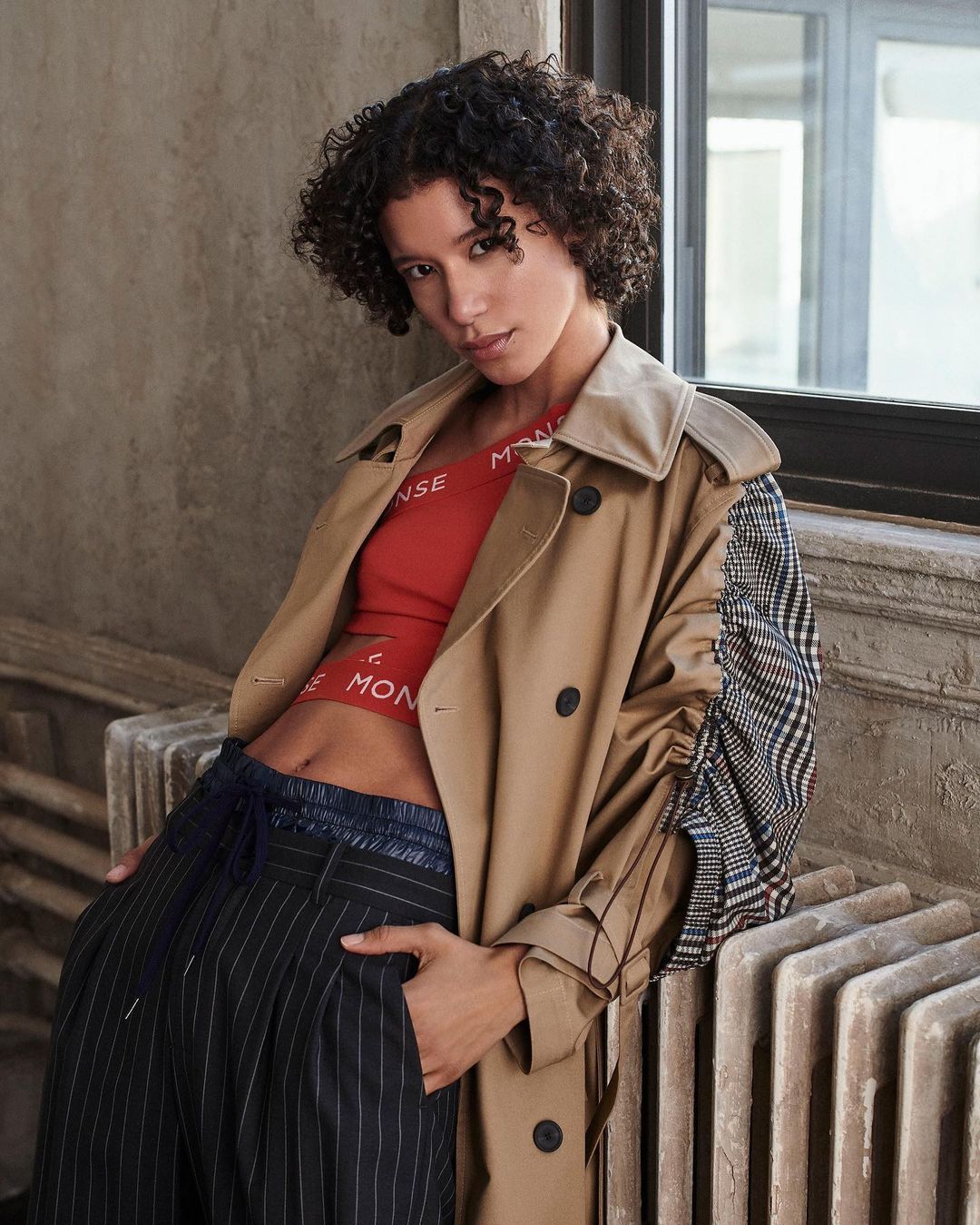 A woman with curly hair poses near a window, wearing a red crop top, pinstripe pants, and a trench coat. Fashion editorial shot.
