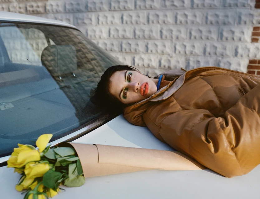 A woman in a brown puffer jacket lies on the hood of a white car next to a bouquet of yellow roses, set against a brick and stone wall.
