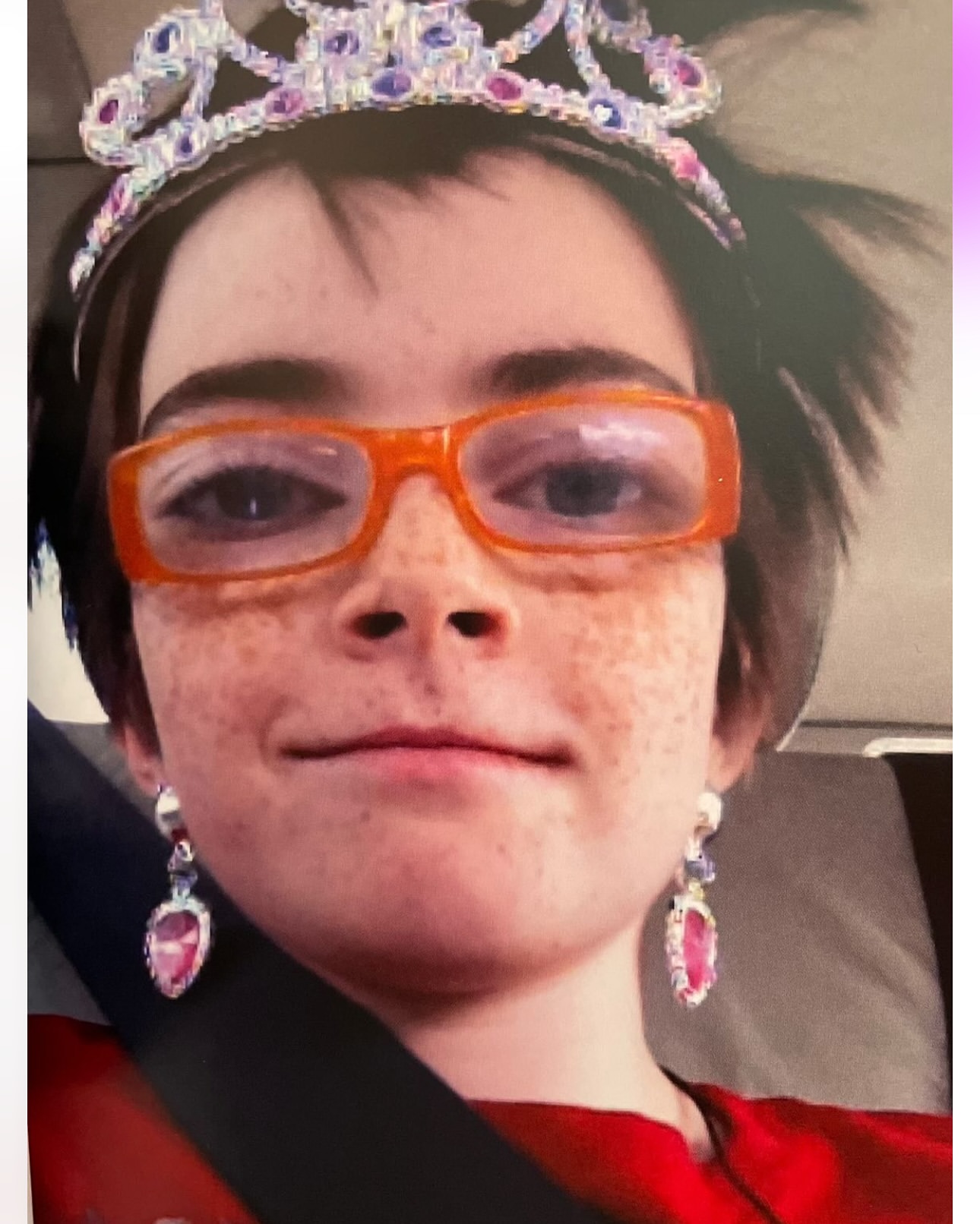 Young girl with a jeweled tiara, orange glasses, and dangling earrings. Inside vehicle with a seatbelt.