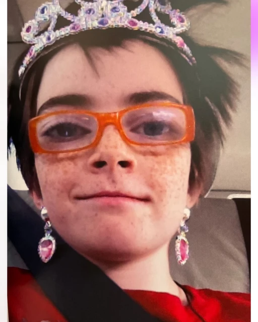 Young girl with a jeweled tiara, orange glasses, and dangling earrings.  Inside vehicle with a seatbelt.