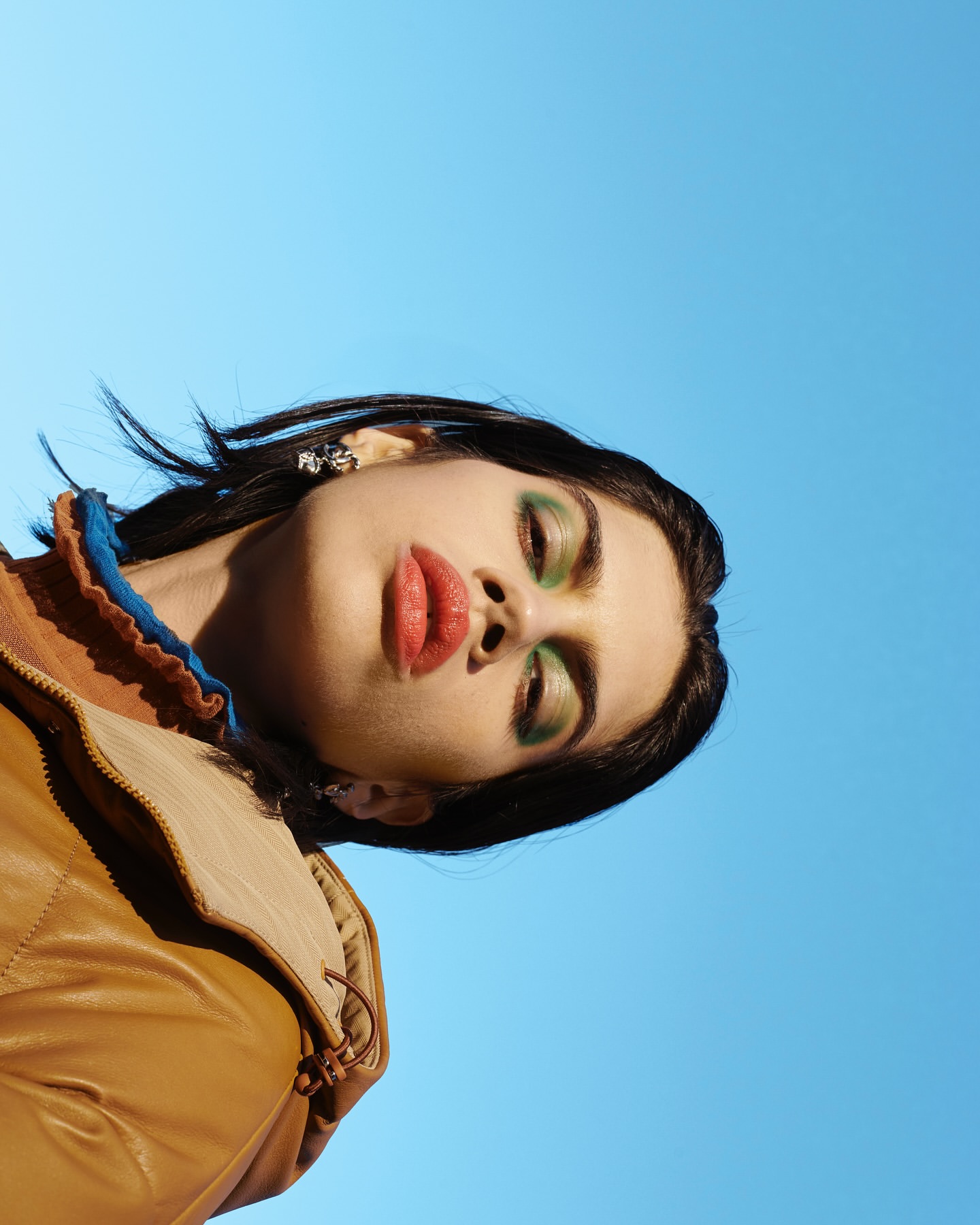 Eye-level shot of a young woman with green eye shadow and a tan jacket, set against a clear blue sky, looking directly at the camera.