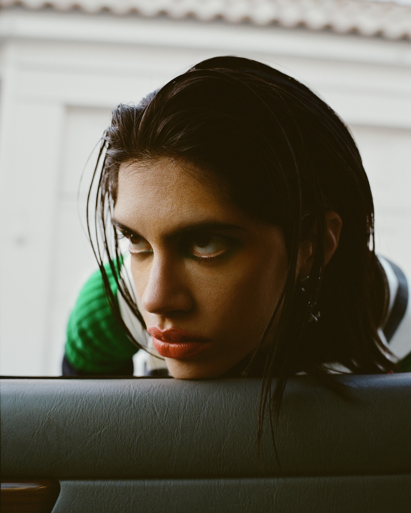A woman with dark hair and fair skin leans against the grey panel of a car door in a close-up portrait with a neutral expression.