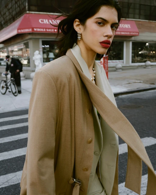 A woman in a tan blazer walks on a city sidewalk in front of a jewelry store, captured in a medium shot with soft, natural lighting.