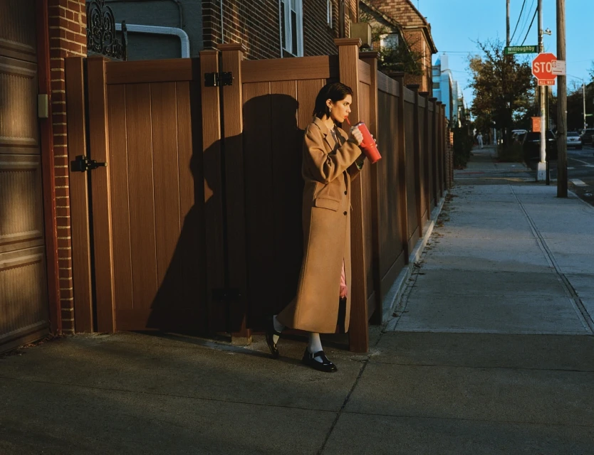 Woman in a tan coat drinks from a red cup while standing on a sidewalk next to a brown fence on a sunny day in an urban environment.