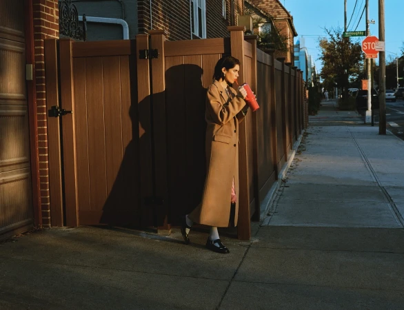 Woman in a tan coat drinks from a red cup while standing on a sidewalk next to a brown fence on a sunny day in an urban environment.