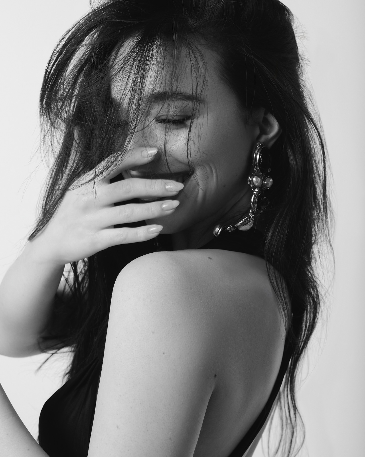 Black and white portrait of a smiling woman with hand covering mouth and statement earring by Tiffany Nicholson