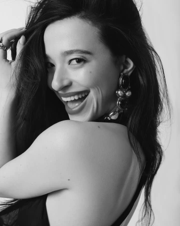 Close-up portrait of a smiling woman with long dark hair, wearing ornate statement earrings against a plain background.