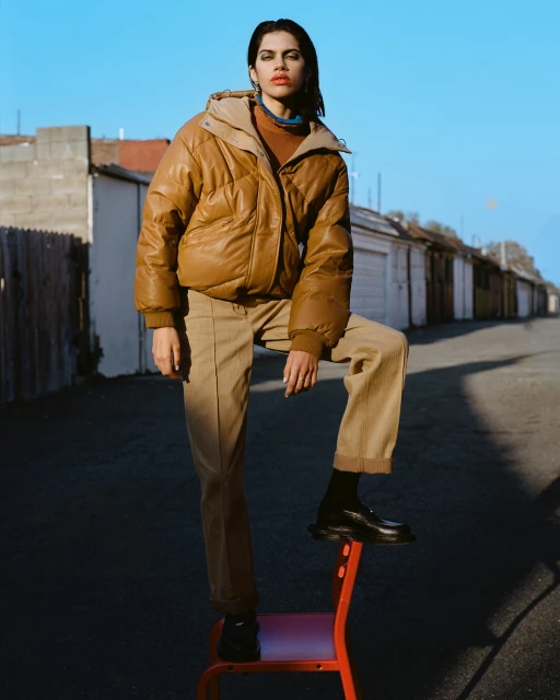 Woman in brown puffer jacket and khaki pants poses on a red chair in an alley with garages under a clear blue sky outdoors.