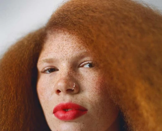 Close-up portrait of a woman with vibrant red hair, heavy freckles, a nose ring, and bold red lips against a neutral background.
