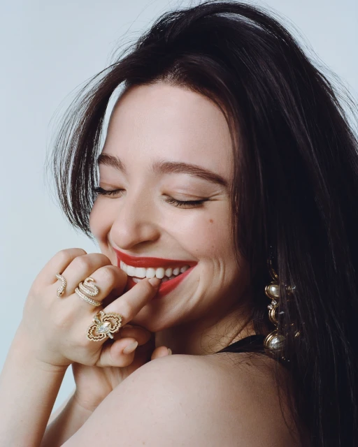 Close-up of a smiling woman with dark hair, wearing red lipstick and multiple gold rings, set against a plain studio backdrop.