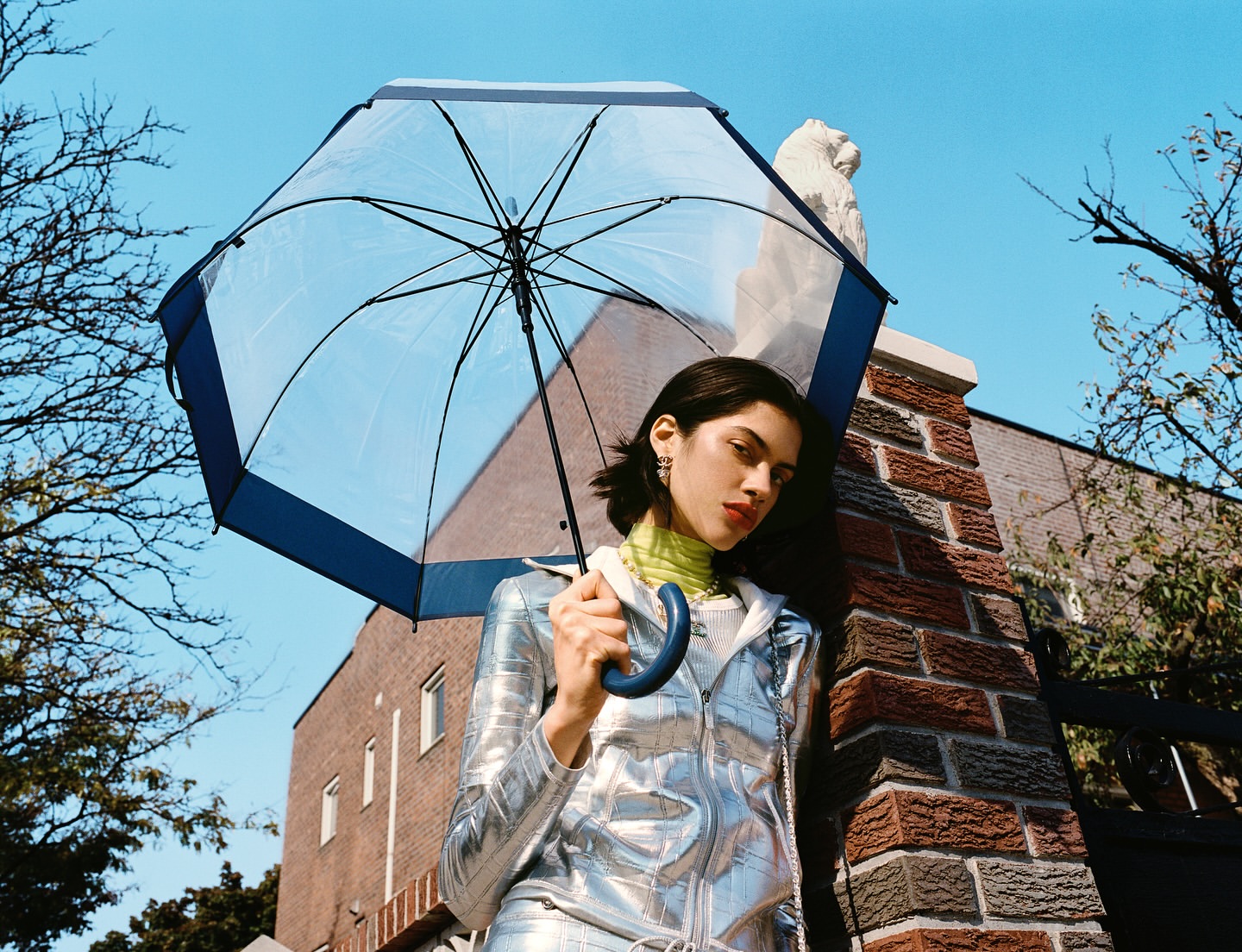 Eye-level shot of a woman in a silver tracksuit holding an umbrella against a blue sky and brick building backdrop on a sunny day.