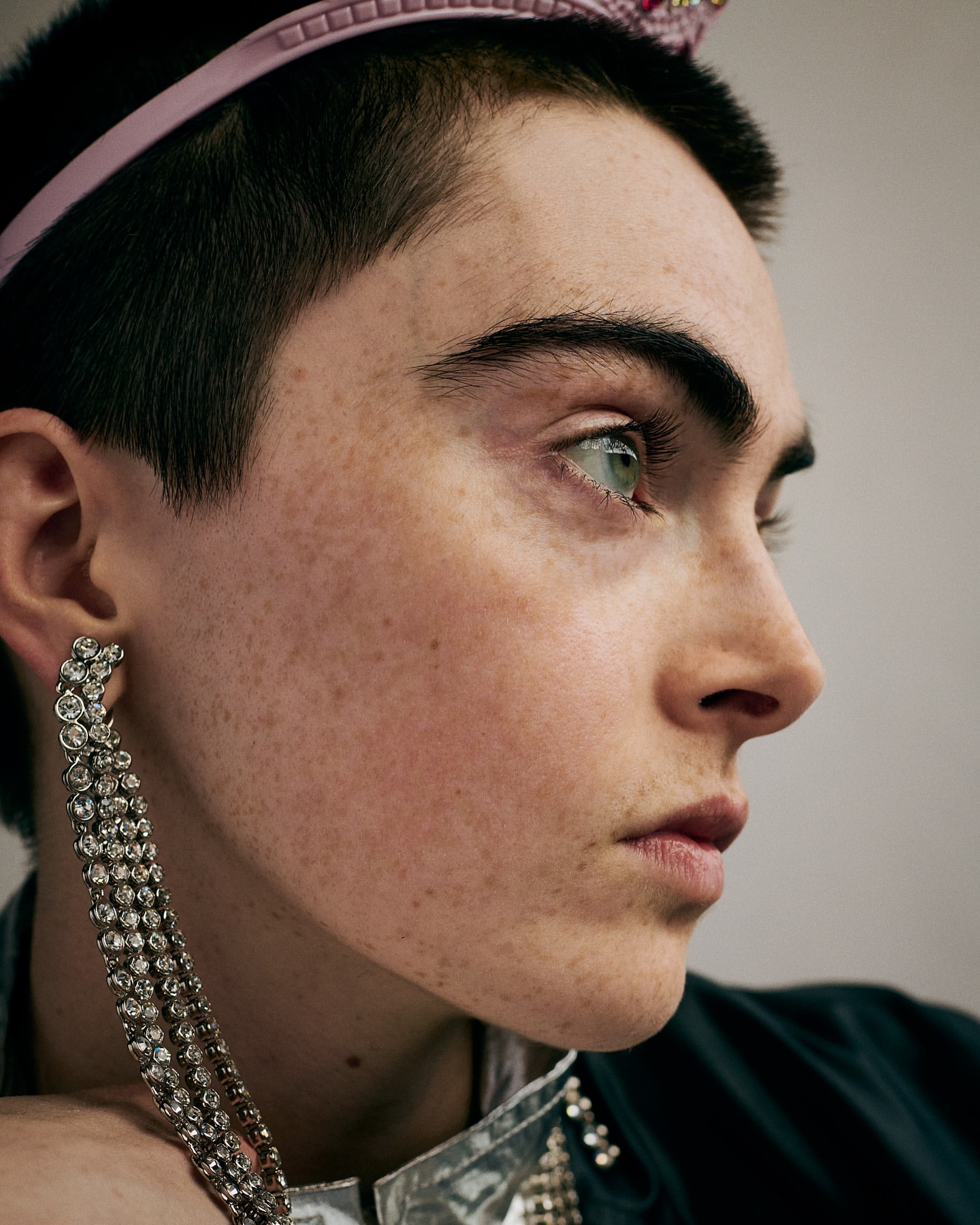 Close-up portrait of a person with short dark hair wearing a pink headband, statement earrings, and an embellished top, looking off-camera.