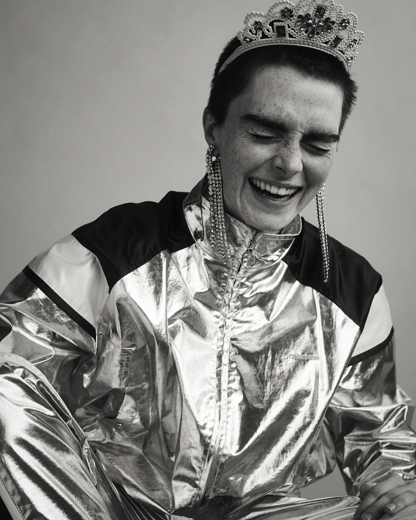 Monochrome portrait of a smiling woman with short hair, wearing a shiny track suit, tiara, and long earrings, indoors.
