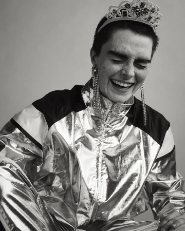 Monochrome portrait of a smiling woman with short hair, wearing a shiny track suit, tiara, and long earrings, indoors.