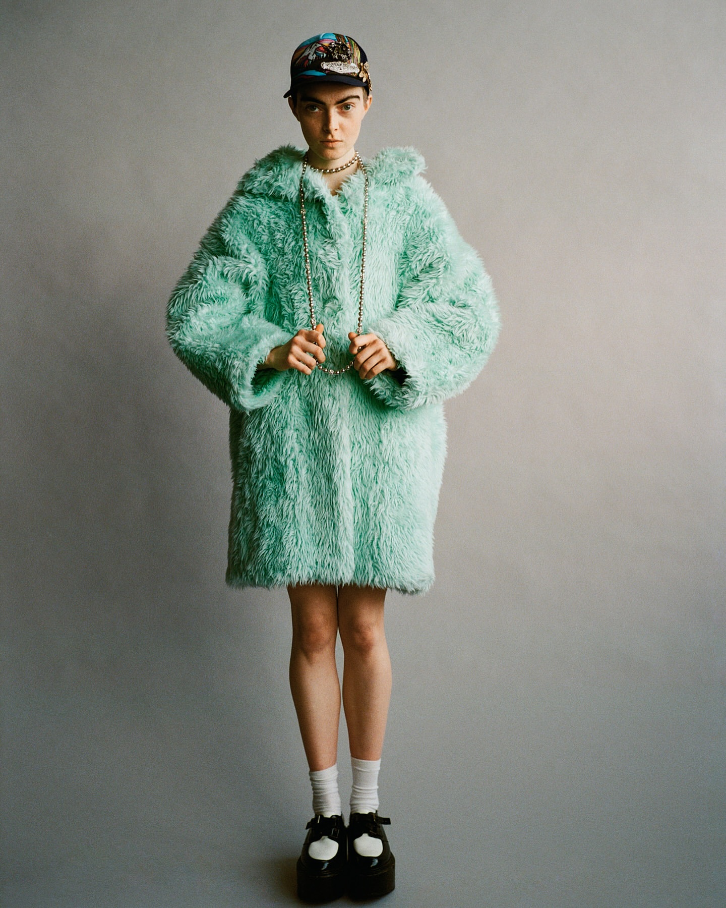 Full shot of a young adult woman in a teal faux-fur coat, cap, necklace, white socks and black Mary Jane platform shoes against a gray background.