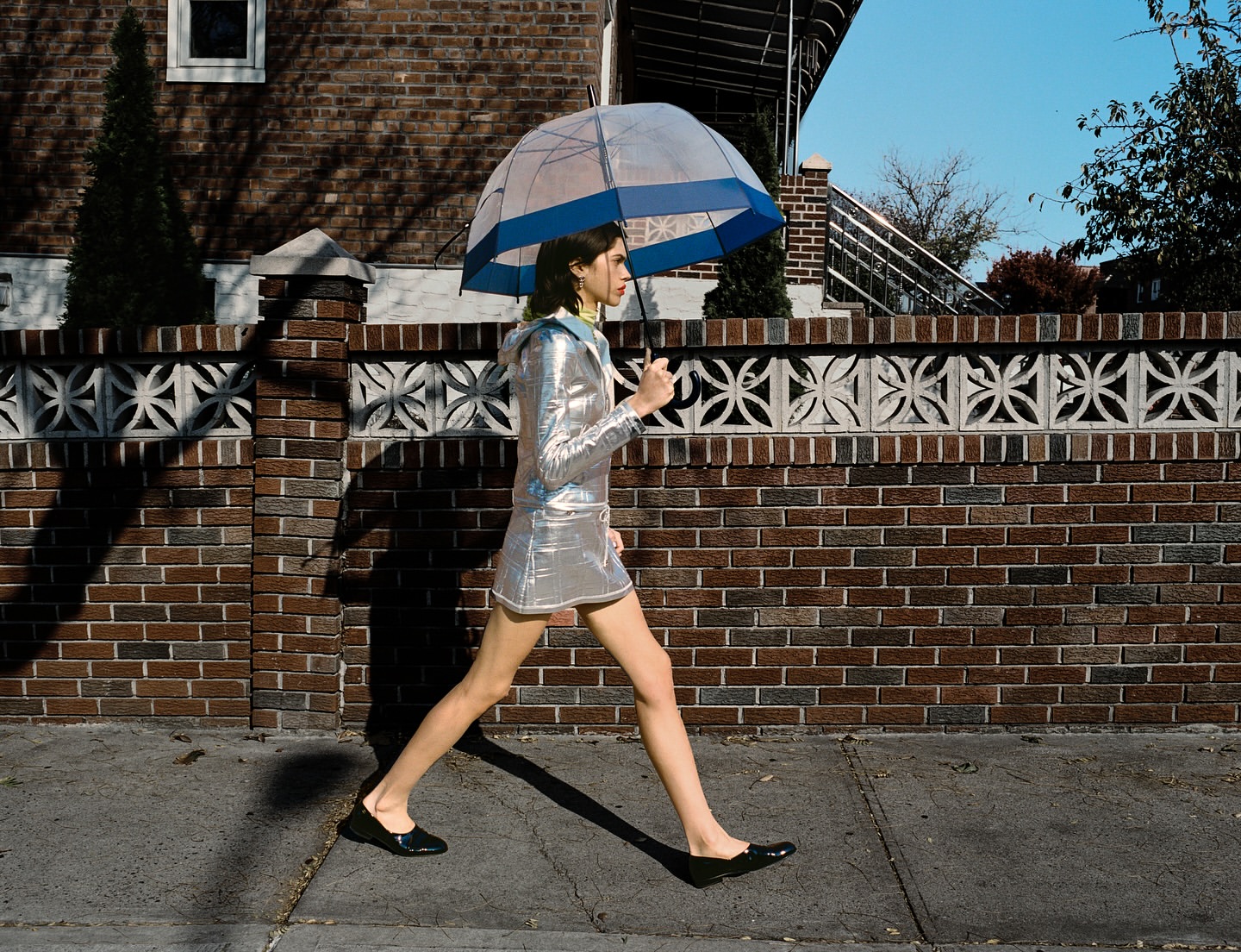 A woman in a silver dress walks on a sidewalk with an umbrella against a brick wall on a sunny day, showcasing an urban fashion style.