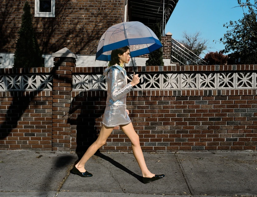 A woman in a silver dress walks on a sidewalk with an umbrella against a brick wall on a sunny day, showcasing an urban fashion style.