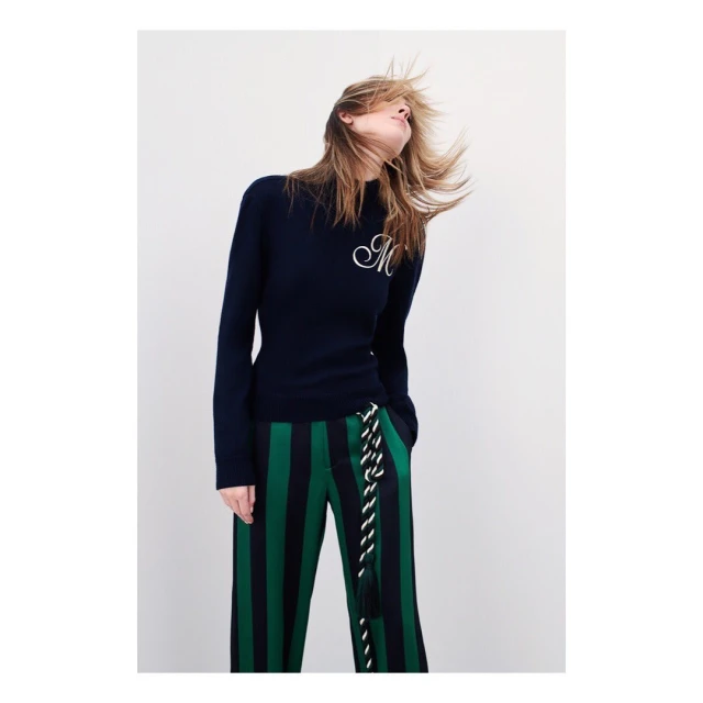 A woman wearing a navy sweater with striped green pants poses against a white background in a fashion editorial style image.