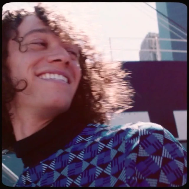 Smiling man with curly hair in geometric patterned shirt with a high-rise building in the background under bright lighting.