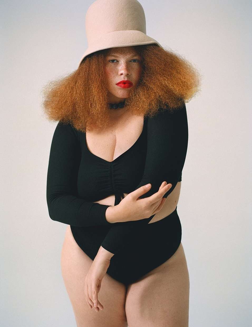 Studio portrait of a red-haired woman wearing a black bodysuit and beige hat against a white backdrop. Beauty, fashion, and studio shot.