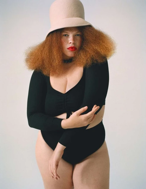 Studio portrait of a red-haired woman wearing a black bodysuit and beige hat against a white backdrop. Beauty, fashion, and studio shot.