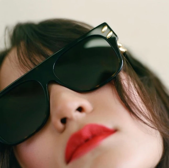 Close-up of a woman with black hair and red lipstick wearing large black sunglasses with gold accents, shot indoors.