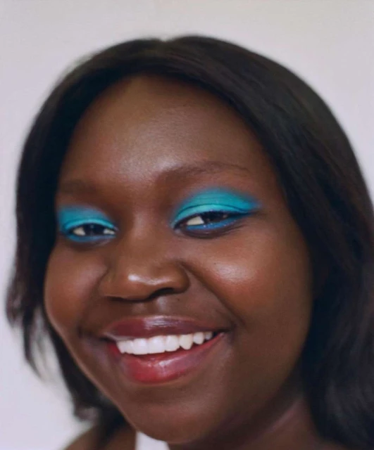 Close-up of a smiling Black woman with dark hair and vibrant turquoise eyeshadow, set against a plain white background in soft, diffused lighting.