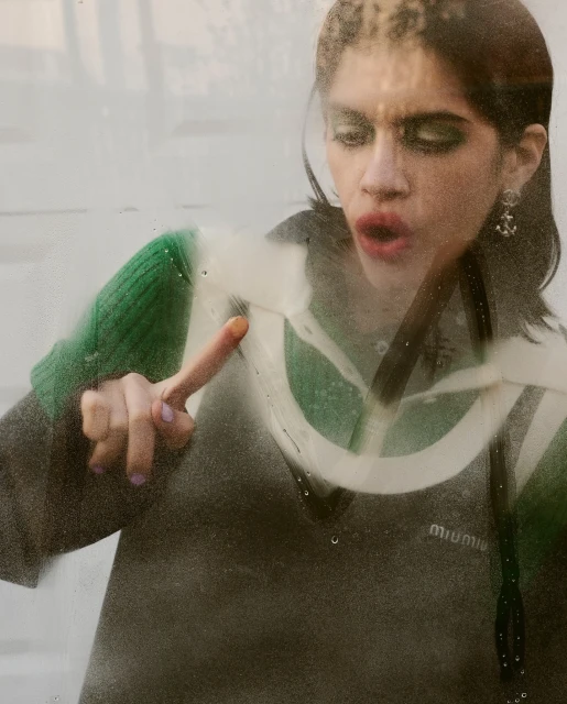 Young woman in fashion sweater draws on steamed glass with finger. Visible makeup, earrings. Indoor, daytime, medium shot. 