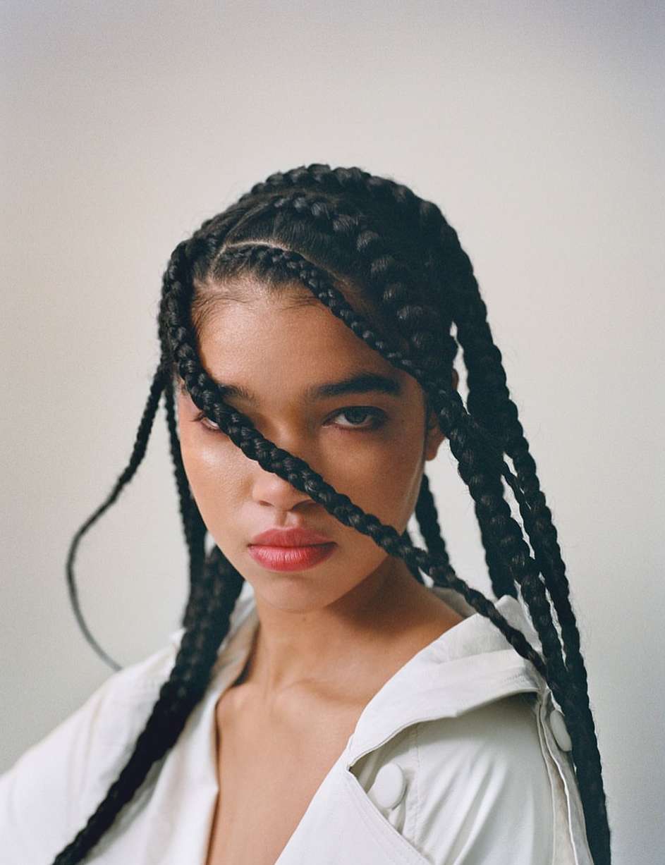 Portrait of a woman with long black box braids wearing an off-white shirt against a plain light background