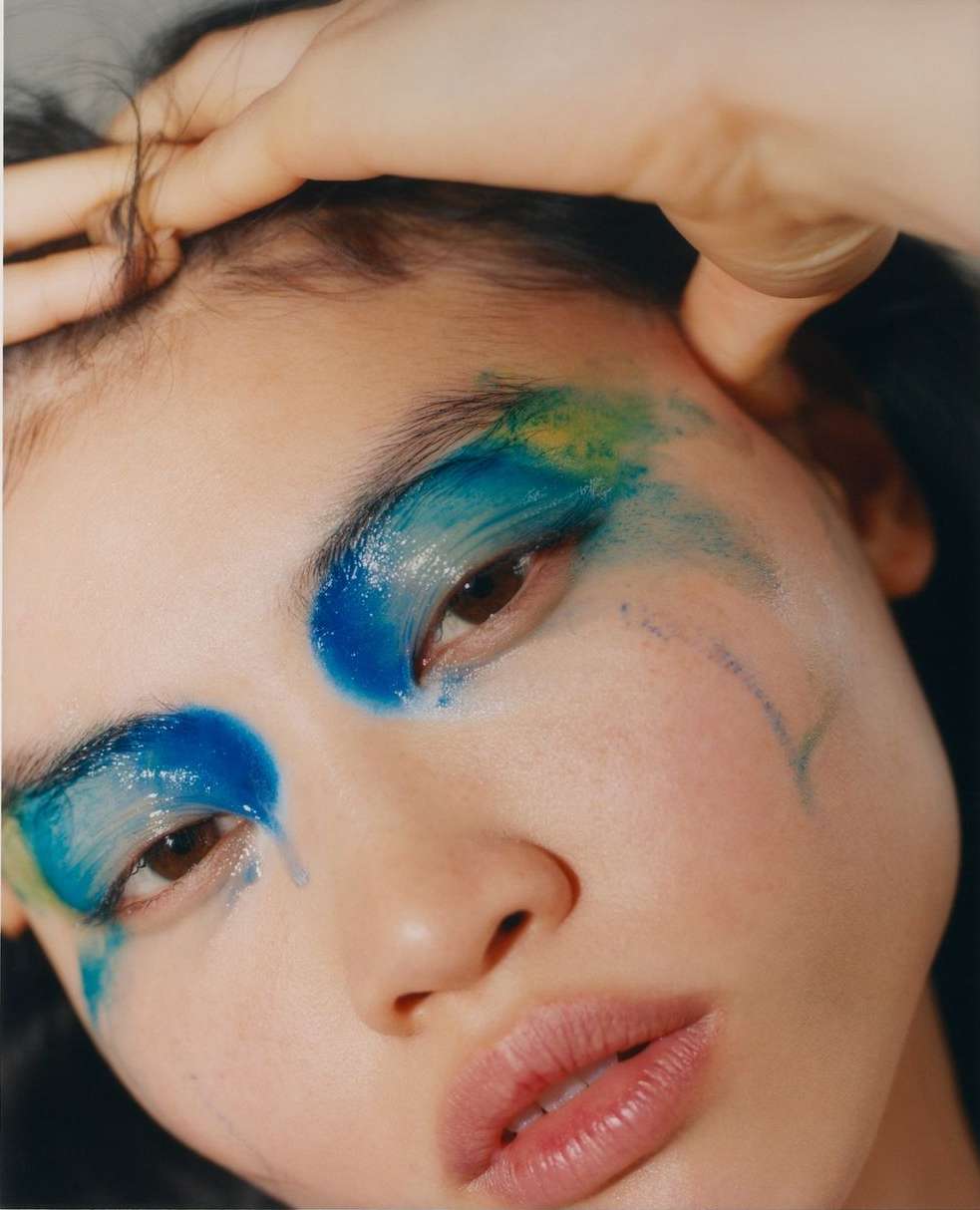 Close-up portrait of a young adult woman with dramatic blue and yellow eye makeup, fair skin, and glossy lips, in a beauty shot.