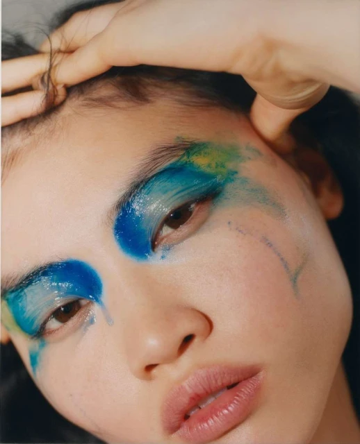 Close-up portrait of a young adult woman with dramatic blue and yellow eye makeup, fair skin, and glossy lips, in a beauty shot.