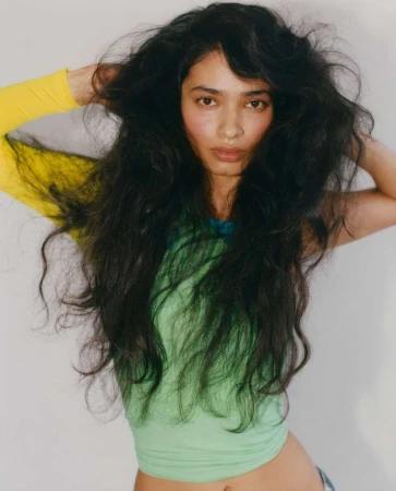 Portrait of a woman with long dark wavy hair holding her hands behind her head wearing a green top