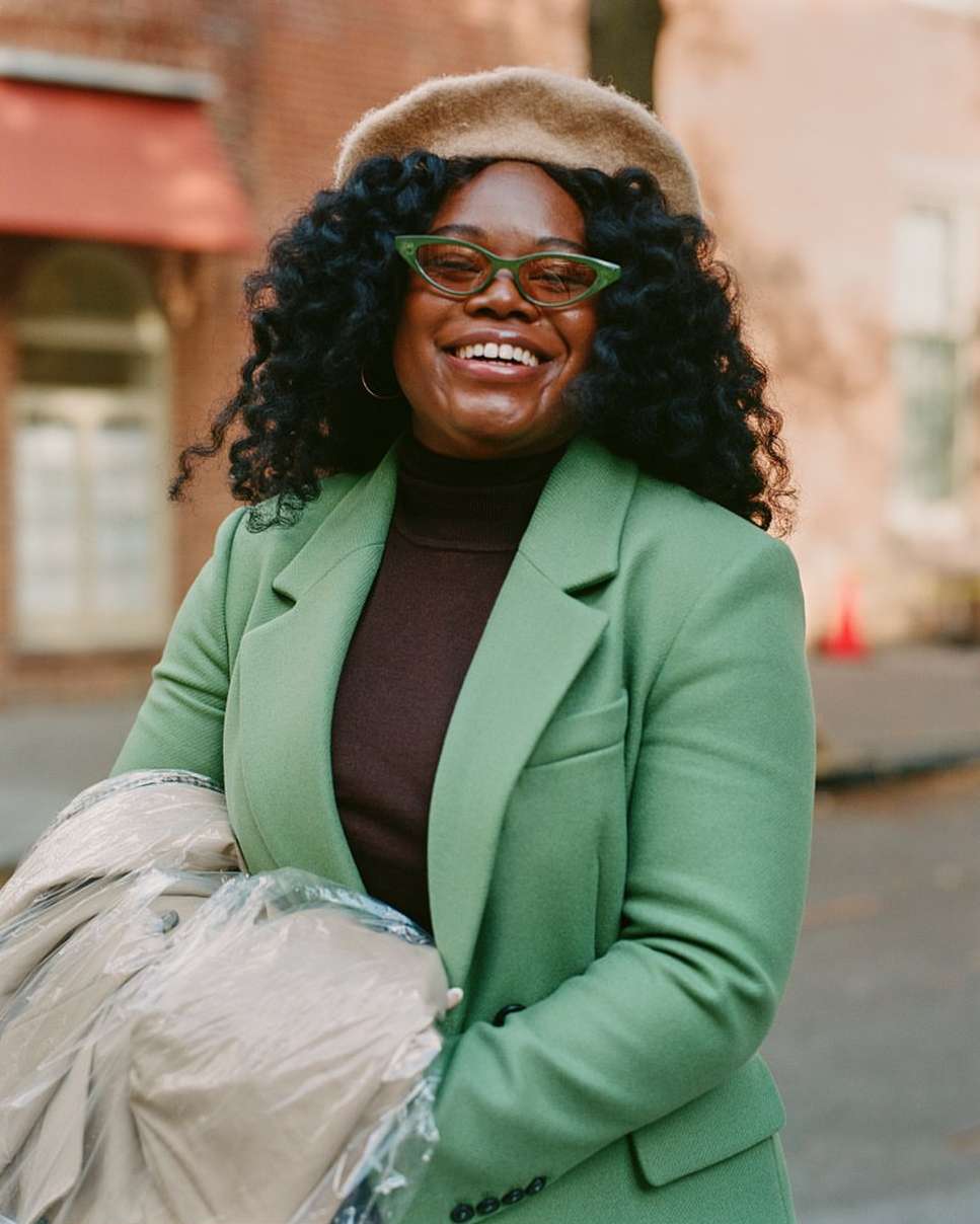 Cheerful woman in a green blazer, brown turtleneck, and beret poses outdoors with a happy expression in medium eye-level shot.