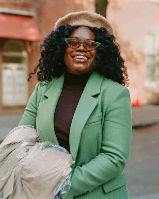 Cheerful woman in a green blazer, brown turtleneck, and beret poses outdoors with a happy expression in medium eye-level shot.