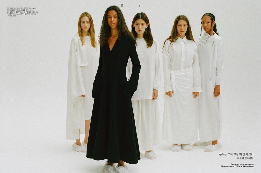 Five fashion models pose in white studio, with one model in a long-sleeved black gown. Models are in matching white slippers, studio lighting.