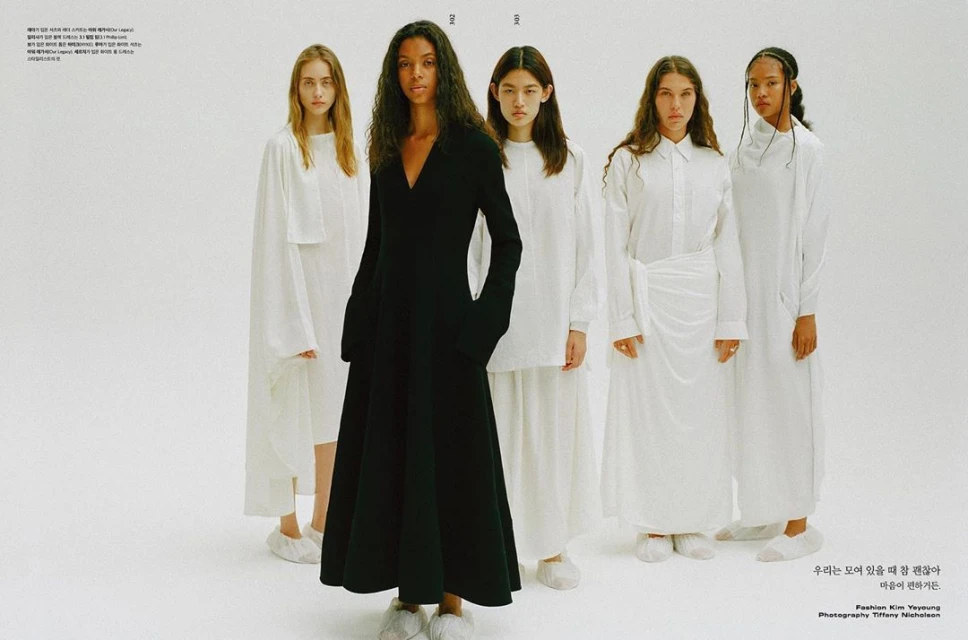 Five fashion models pose in white studio, with one model in a long-sleeved black gown. Models are in matching white slippers, studio lighting.