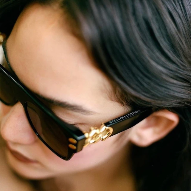 Person wearing Stella McCartney sunglasses, close-up studio shot. Dark hair frames the face, emphasizing the gold detailing on the eyewear.